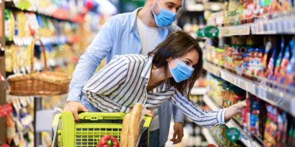 Shopping Together. Portrait of millennial family wearing protective surgical masks standing in supermarket mall with cart, buying food and groceries. Couple choosing healthy products and nutrition
